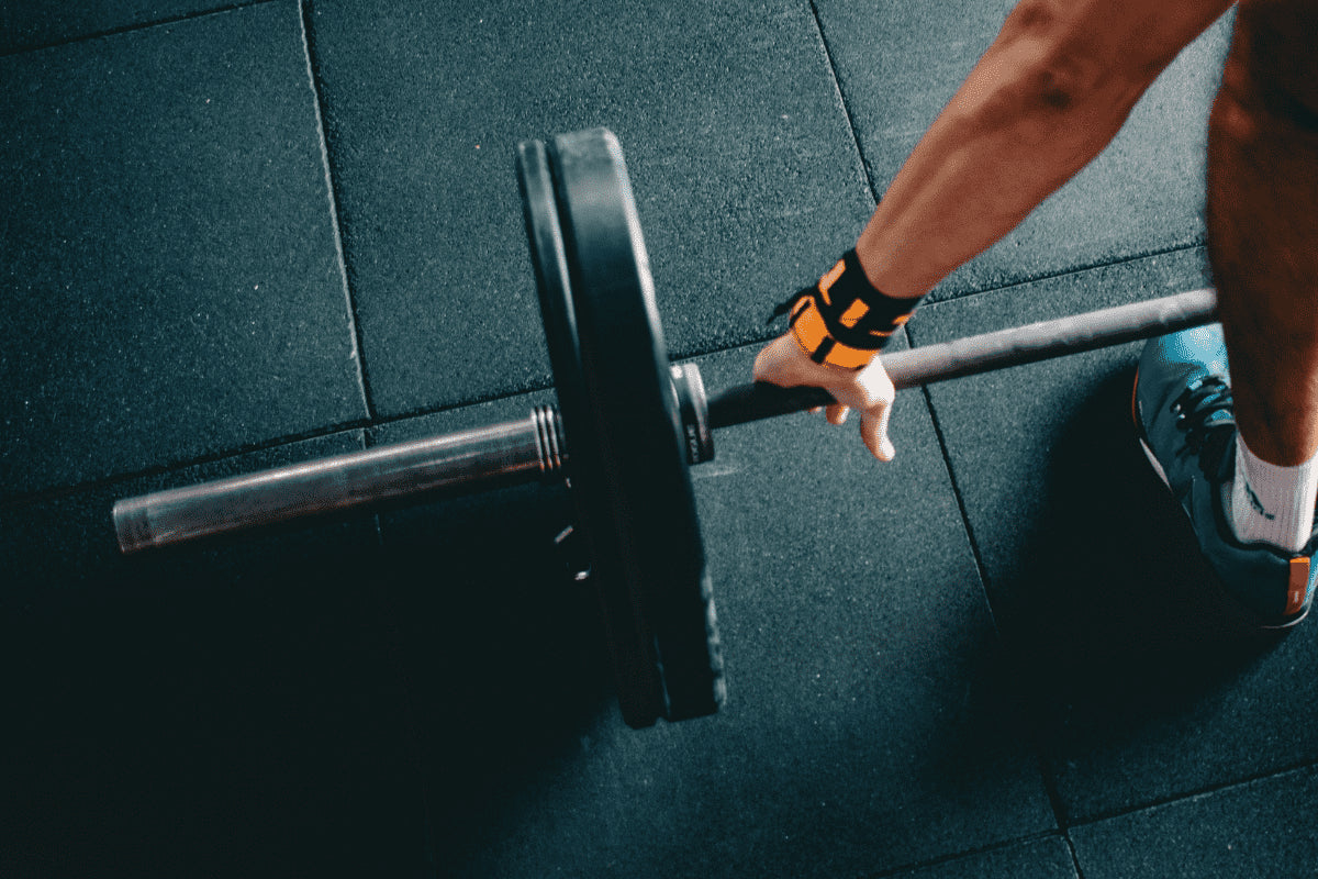 close up photo of a barbell with bumper plates on a rubber gym floor. you can see a mans handing grasping the bar